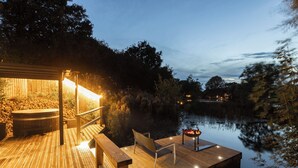 Um deck de madeira com hidromassagem, área de estar e lareira à beira de um lago tranquilo ao entardecer.