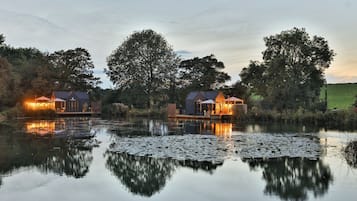 A lakeside property with two illuminated buildings, a calm lake with lily pads, and surrounding trees.