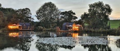Uma propriedade à beira de um lago com dois edifícios iluminados, um lago calmo com vitórias-régias e árvores ao redor.