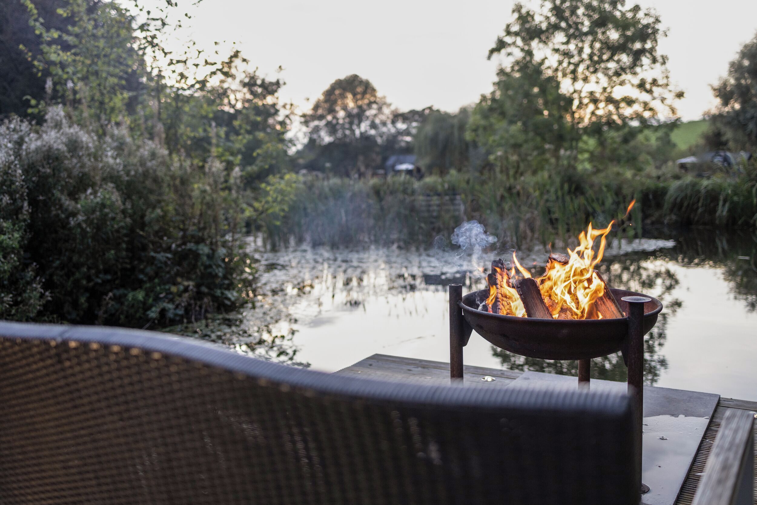 A fire pit with flames on a deck overlooking a calm body of water, surrounded by lush greenery.