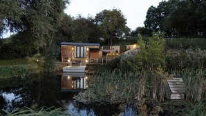 Uma cabana de madeira moderna à beira de um lago tranquilo, rodeada por vegetação exuberante e árvores.