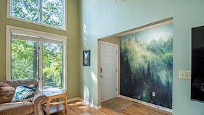 A spacious room with a large window, a cozy leather sofa, a small table, and a forest-themed wall mural. - Private Hot Tub and Resort Amenities Included! (Massanutten)