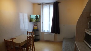 Una stanza piccola e funzionale, con un tavolo, due sedie, una televisione a muro, una finestra con tende e un termosifone.