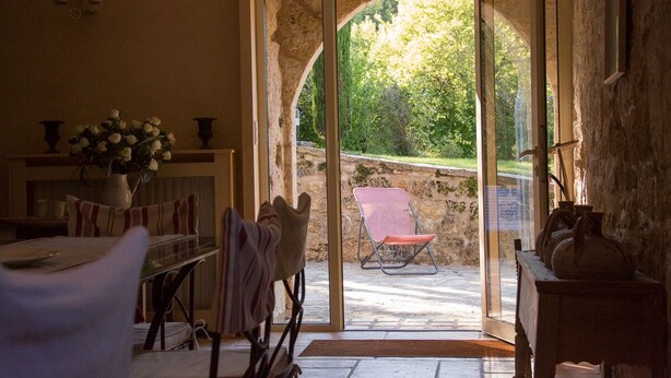 A room with a table, chairs, a vase of flowers, and a view through an arched doorway to an outdoor area with a chair and a table.