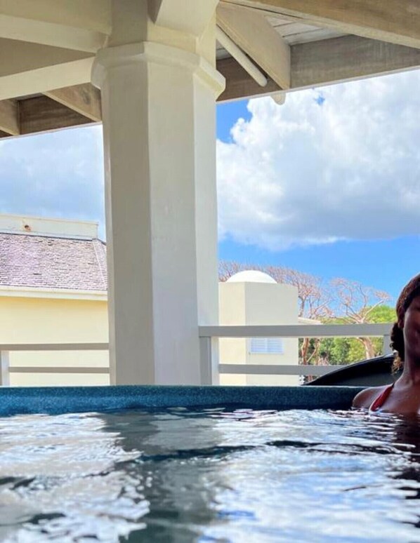 Eine Person in einer Hot-Whirl-Badewanne unter einem Überdachung mit Blick auf Gebäude und Bäume.