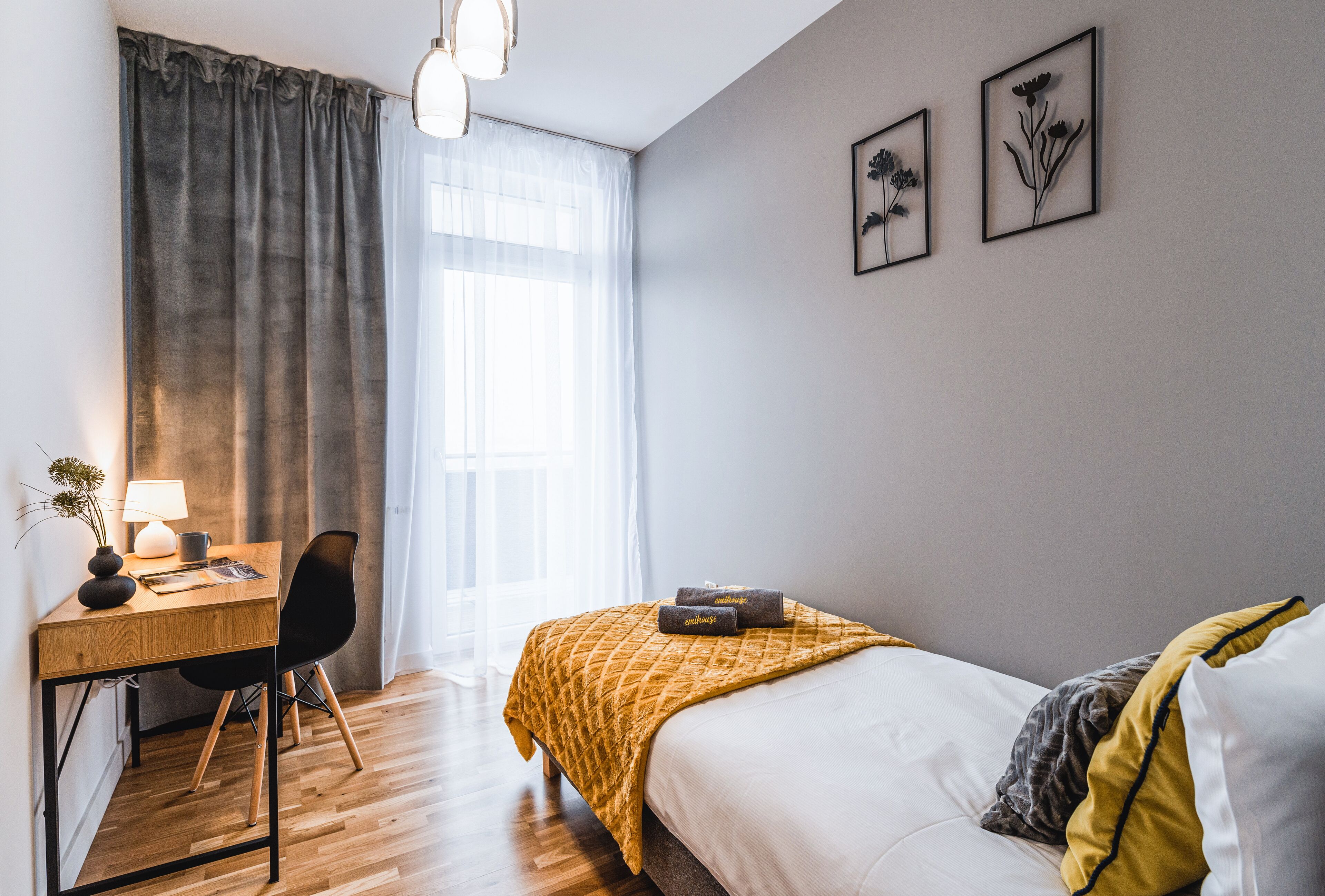 Hotellihuoneessa on puinen työpöytä, tuoli, keltainen peitto sängyllä sekä kaksi kehystettyä, kukkateemalla varustettua taideteosta seinällä.