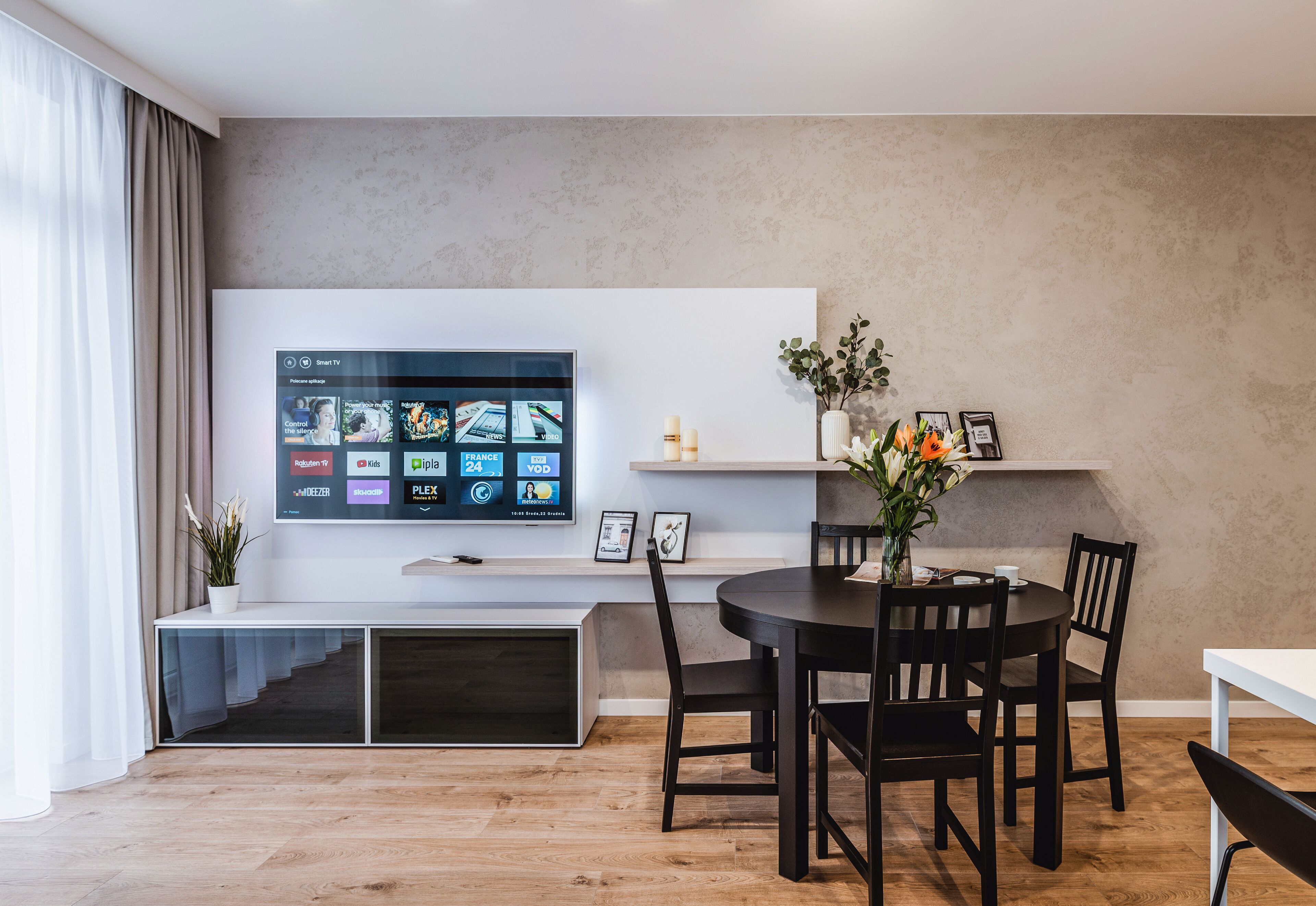 Una sala de estar moderna con un televisor de pantalla plana montado en la pared, una mesa de comedor con sillas y un estante pequeño con objetos decorativos.