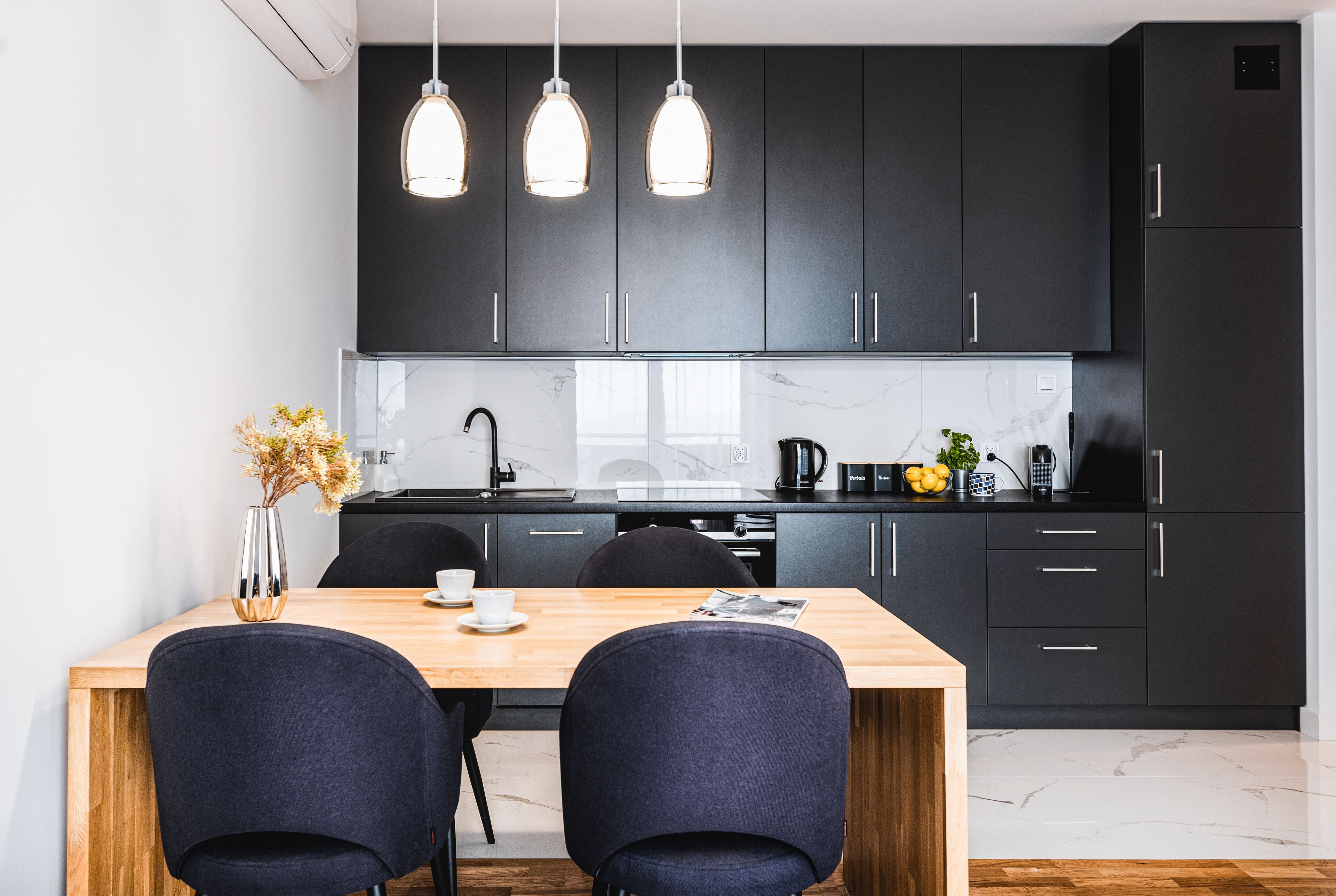 A modern kitchen with dark cabinets, a wooden dining table, and pendant lights.