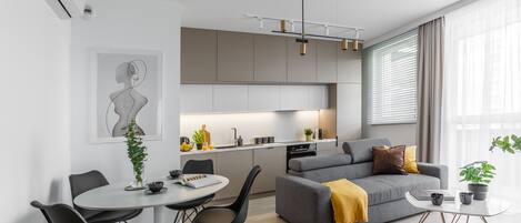 Una sala de estar moderna con un sofá gris, una mesa de comedor redonda y una cocina con armarios grises y encimera blanca.