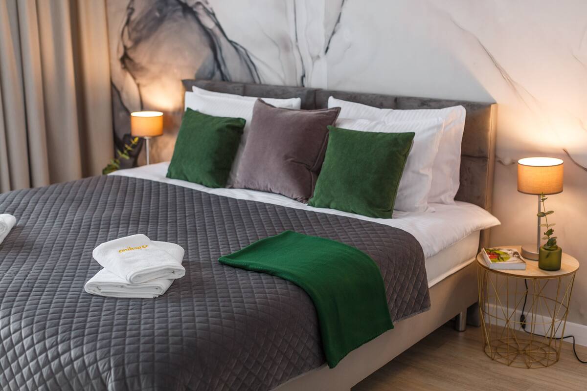 Een net opgemaakt bed met een grijze sprei, groene en bruine kussens, en een gevouwen witte handdoek ernaast. Daarbij staat een klein rond tafeltje met een lamp en een plantje.