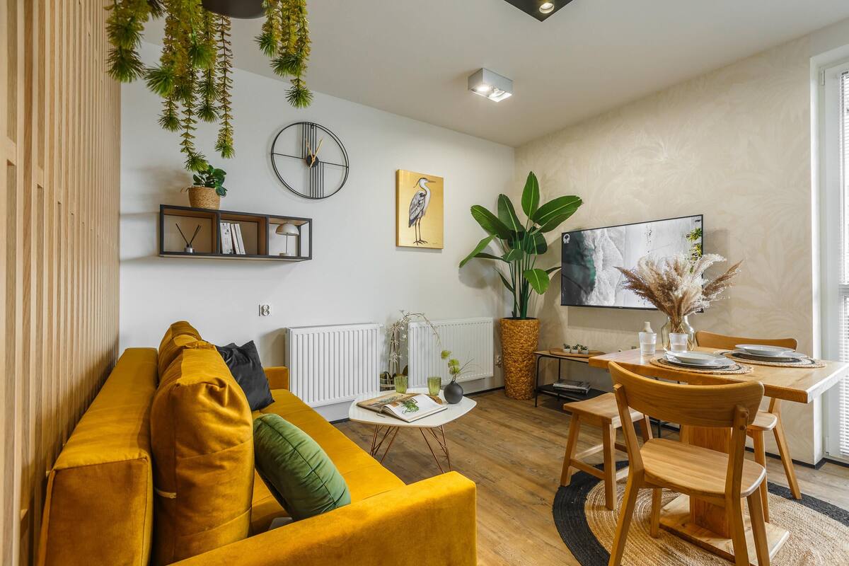 Een moderne woonkamer met een gele bank, een eettafel met stoelen, een grote plant en een hangklok.