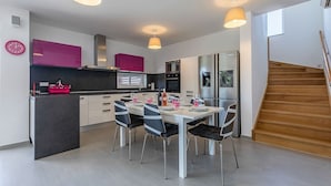 A modern kitchen with a dining area, featuring a central island, pendant lights, and a staircase leading to an upper level.