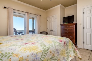 A bedroom with a floral bedspread, a dresser with a television, and a view through the window.