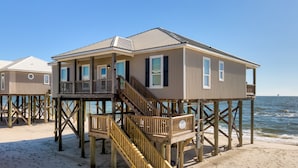 Une maison de plage en bois, dotée d’un balcon et d’un escalier, construite sur pilotis au-dessus du sable.