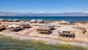 Vue aérienne d’une zone côtière avec de nombreuses constructions en bois sur pilotis, probablement des maisons de plage, offrant une vue dégagée sur l’océan et une plage de sable au premier plan.