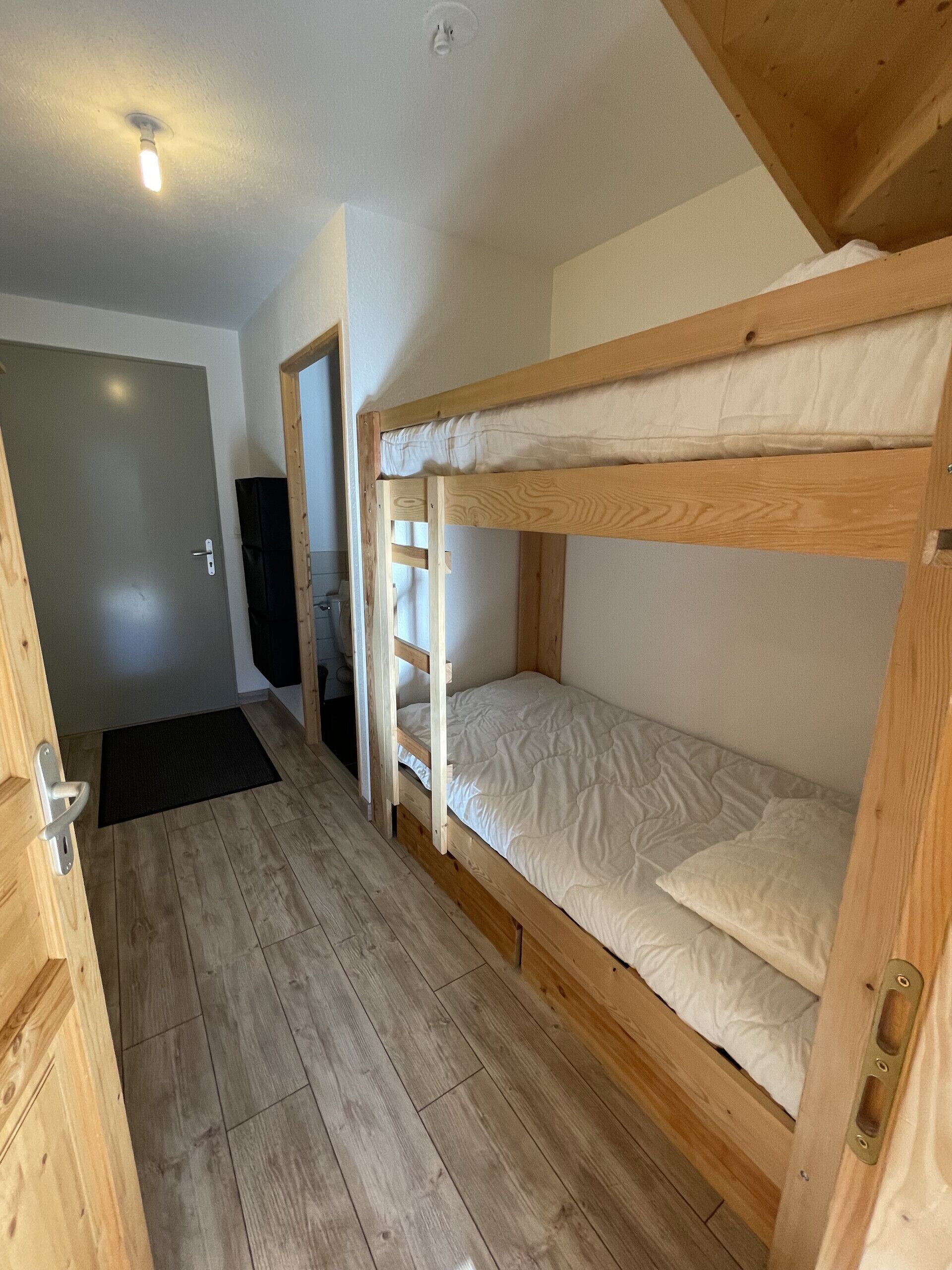 A room with two bunk beds, wooden flooring, a door, and a ceiling light.