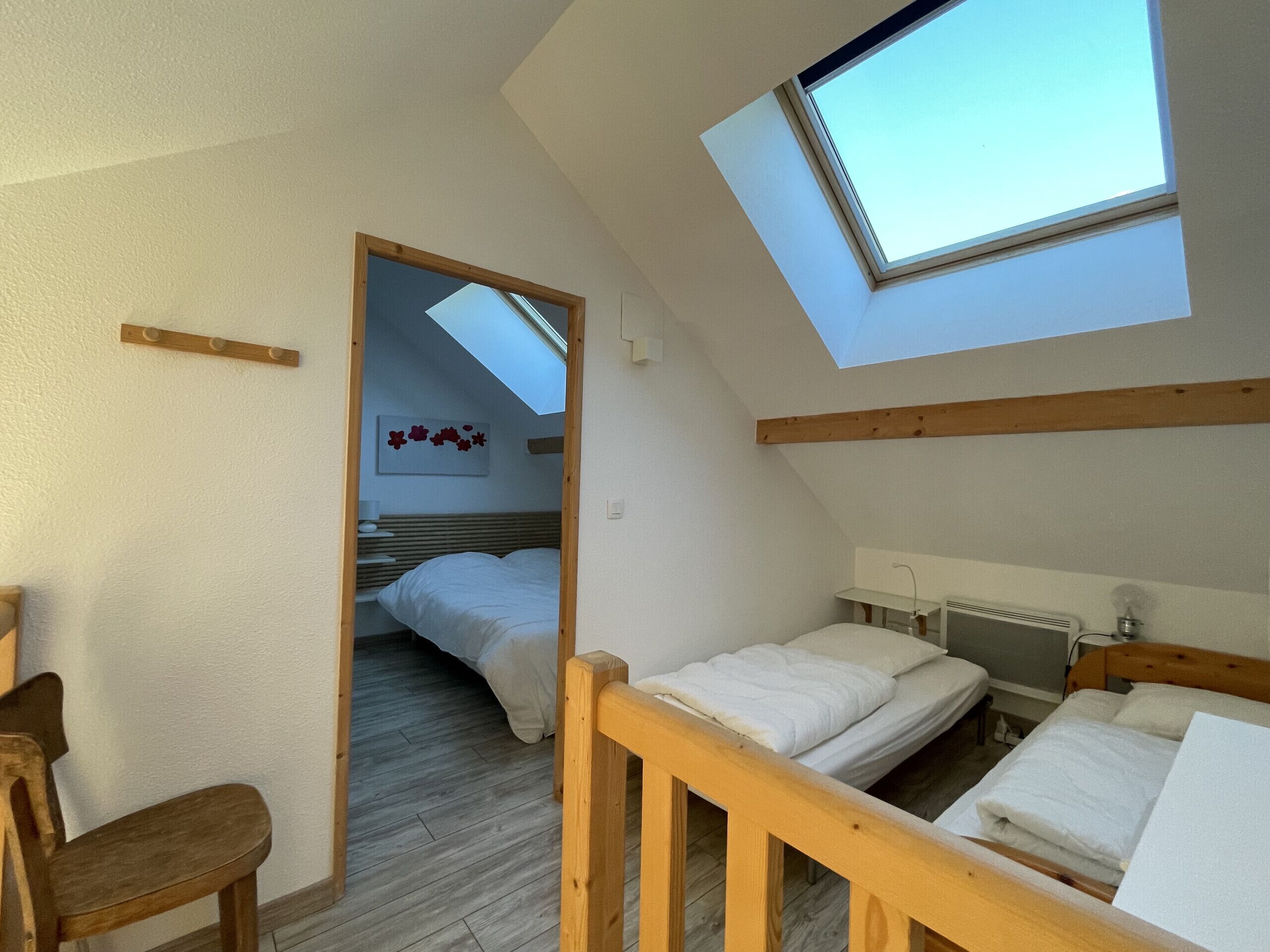 Une chambre avec une lucarne, des poutres apparentes, un lit, une chaise et une table de chevet.
