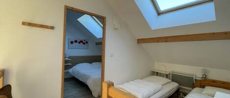 Une chambre avec une lucarne, des poutres apparentes, un lit, une chaise et une table de chevet.