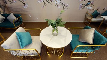 A room with a round white table, two teal cushioned chairs with gold frames, and a vase with green plants.