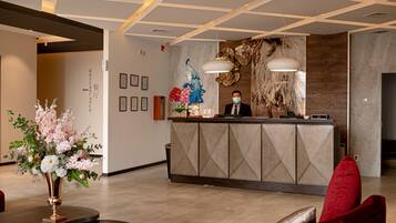 A hotel reception area with a modern design, featuring a reception desk, a vase of flowers, and a seating area with red and brown upholstered chairs.