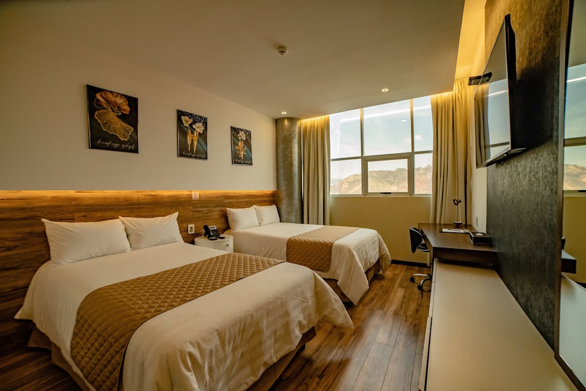 A hotel room with two beds, a desk, a TV, and a window with a view of the mountains.