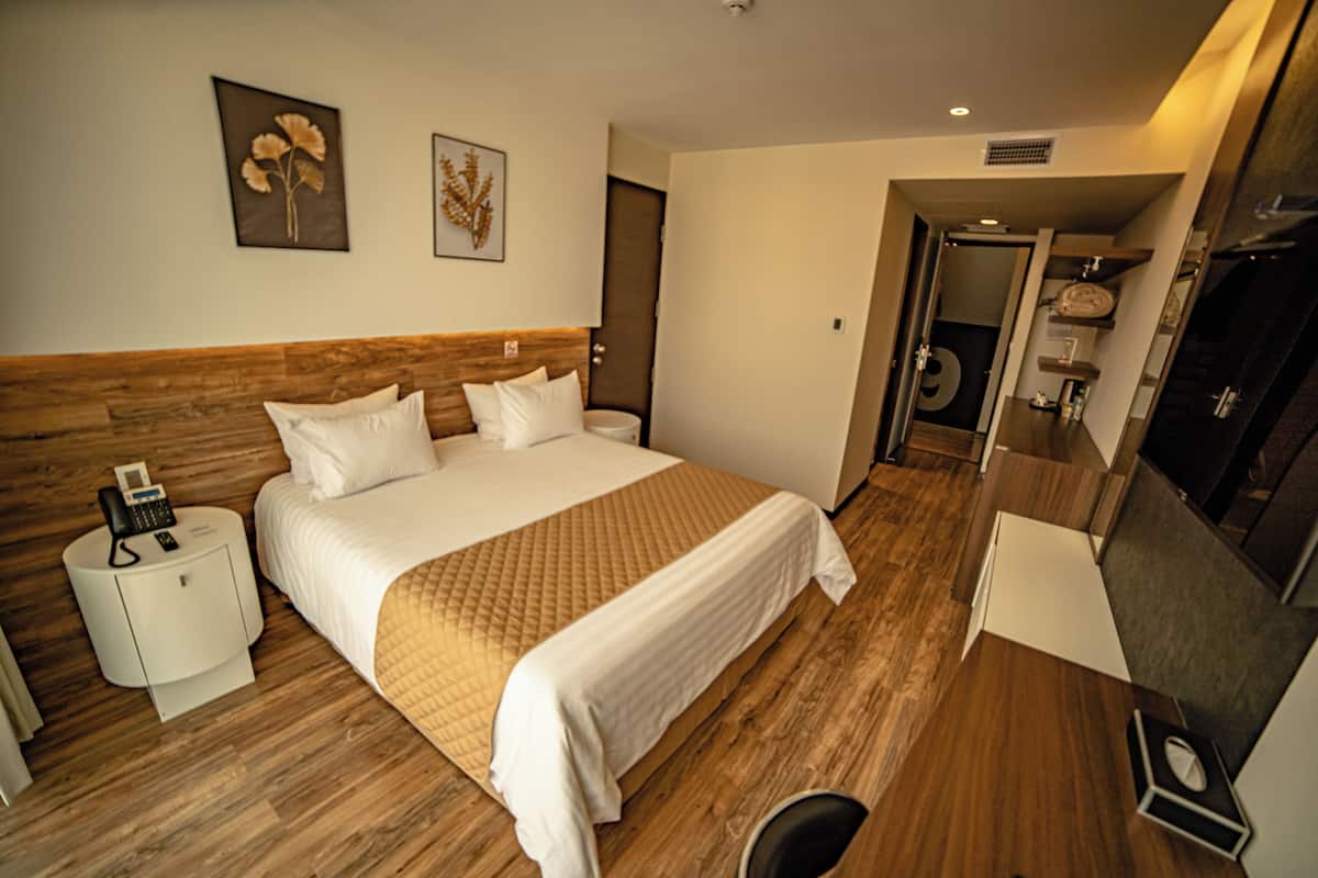 A hotel room with a wooden headboard, a bed with white bedding, a bedside table with a phone, and a small desk with a lamp.