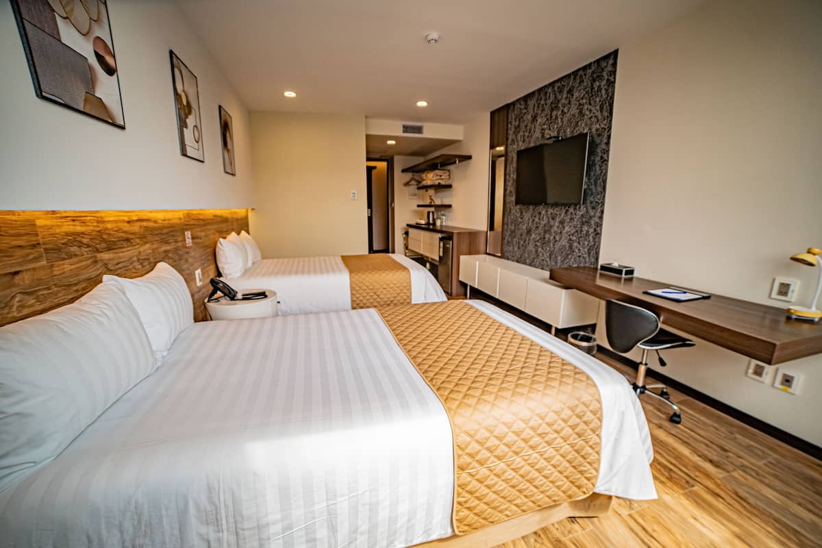 A hotel room with two beds, a desk, a chair, a TV, and a small kitchenette.