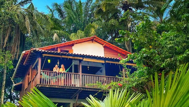 A wooden house with a balcony, surrounded by lush greenery and palm trees. - POUSADA HORIZONTE AZUL (Cairu)