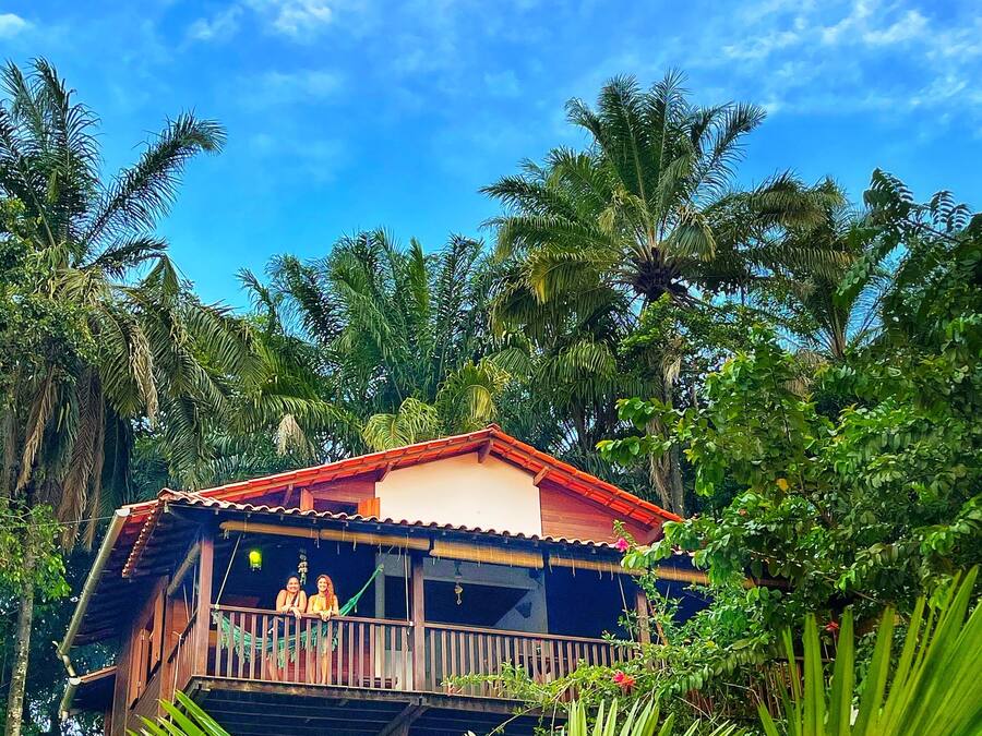 A wooden house with a balcony, surrounded by lush greenery and palm trees.