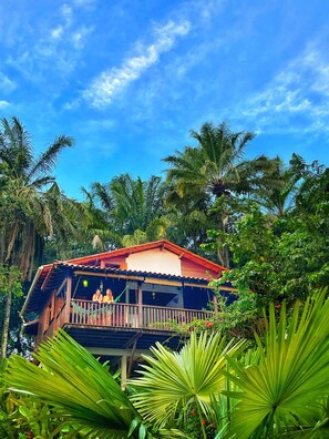 A wooden house with a balcony, surrounded by lush greenery and palm trees. - POUSADA HORIZONTE AZUL (Cairu)
