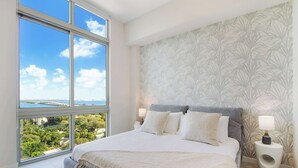 A bedroom with a large window, a bed with white bedding, a grey headboard, and a patterned wallpaper. - Penthouse W/Bayviews! 2 Bdrm Condo In Miami Design (Miami)