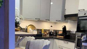 A modern kitchen with white cabinets, a black countertop, and a built-in oven. There are two white bar stools with gold legs. - Superb sauna jacuzzi apartment Luxury&Spa (Herblay)