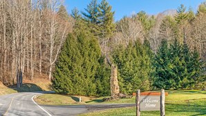 Un chemin sinueux menant à une propriété où est affiché un panneau « Bienvenue chez vous », le tout entouré d’arbres denses et d’une construction en pierre.
