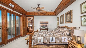 Une chambre avec un grand lit, une tête de lit en bois et un ventilateur de plafond. Des tableaux encadrés ornent le mur, et une fenêtre offre une vue sur l’extérieur.