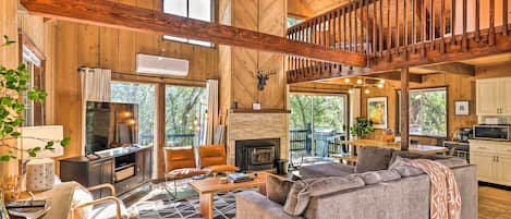 A spacious living room with a fireplace, wooden beams, and a large window overlooking a deck.