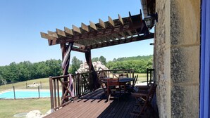 Una terraza de madera con pérgola, mobiliario exterior y vista a una piscina.