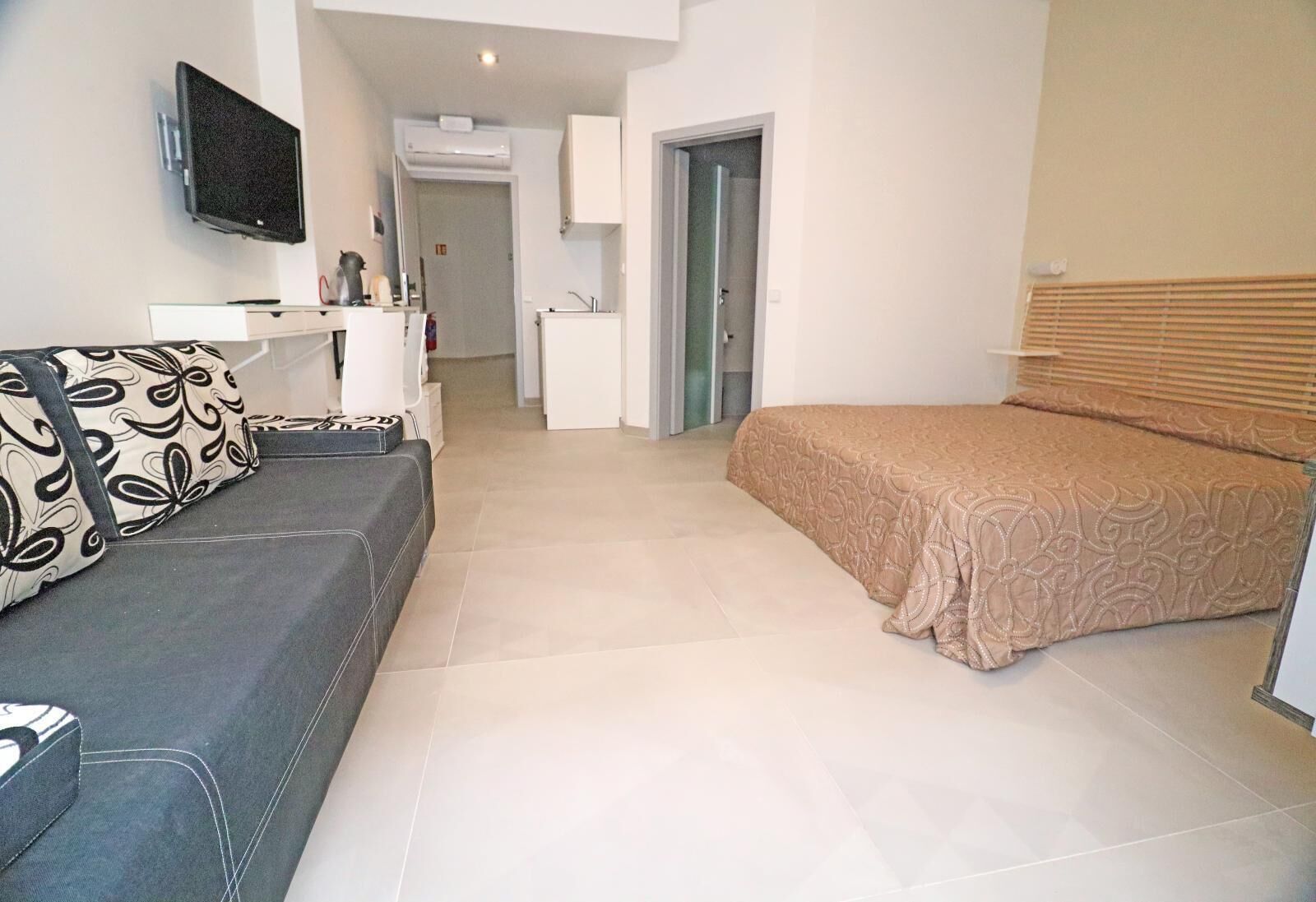 Une chambre d’hôtel moderne avec un canapé gris, un lit recouvert d’une courtepointe à motifs, un téléviseur à écran plat fixé au mur et un petit coin cuisine.