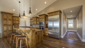A spacious kitchen with wooden cabinets, a stainless steel refrigerator, and a central island with bar stools. - Point Break at Agate Beach, Sleeps 12, Oceanfront, Loft, Game Room, Hot Tub, Beach Access, BBQ (Newport)
