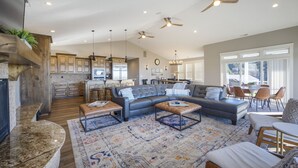 A spacious living area with a large sectional sofa, a coffee table, and a fireplace. There is a kitchen in the background with wooden cabinets and a dining area with chairs. - Point Break at Agate Beach, Sleeps 12, Oceanfront, Loft, Game Room, Hot Tub, Beach Access, BBQ (Newport)