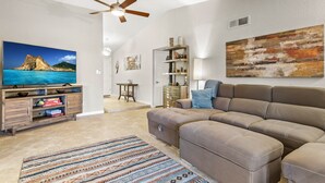 A living room with a sectional sofa, a flat-screen TV, a ceiling fan, and a large abstract painting.