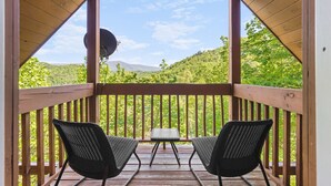 Una terraza de madera con dos sillas de mimbre negro y una mesita, con vistas a un paisaje verde y exuberante con colinas al fondo.