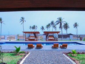 A pool area with two wooden beds and lounge chairs, surrounded by palm trees and a clear view of the ocean. - Impeccable 3-bed House in Prampram Accra (Prampram)