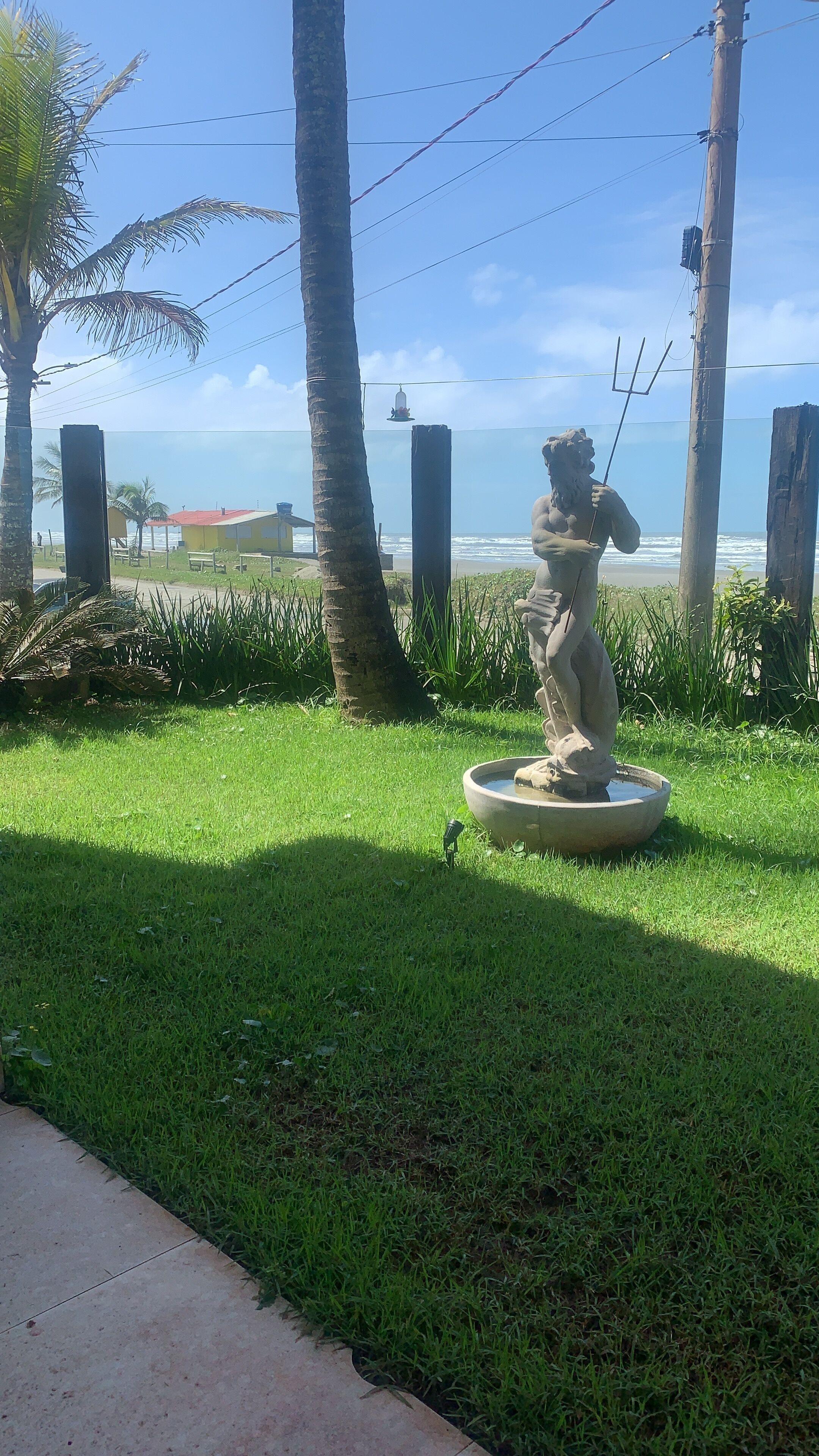 Une statue représentant une figure tenant un trident, entourée d’herbe et de palmiers, avec l’océan en toile de fond.