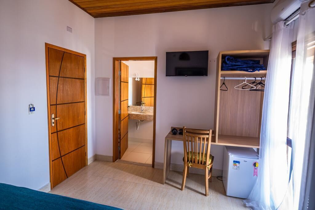 Habitación compacta con puerta de madera, una mesita con una silla, televisor montado en la pared y armario con espacio para colgar.
