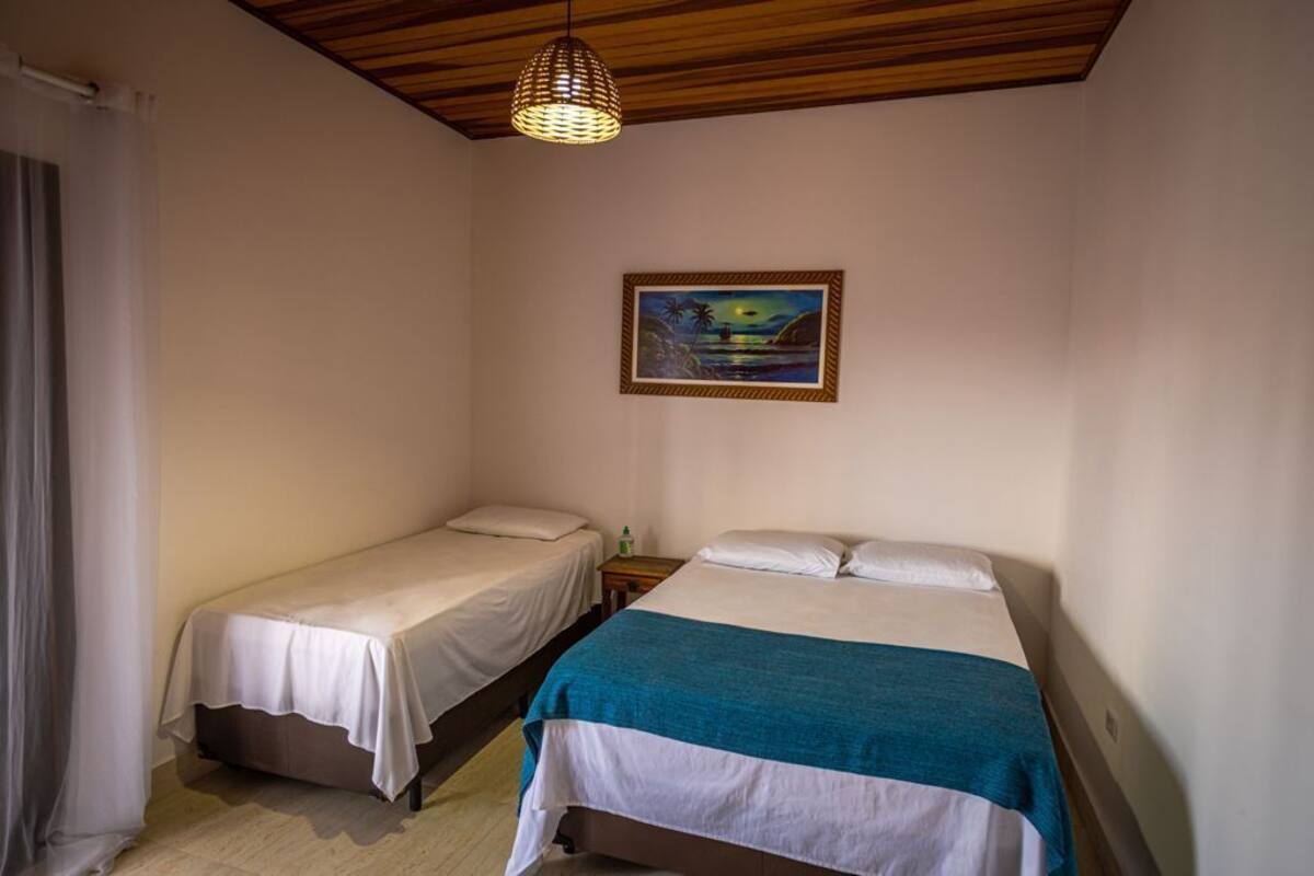 A room with two single beds, a wooden ceiling, a painting on the wall, and a hanging light fixture.