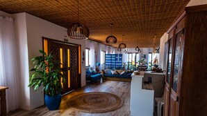 Een ruime woonkamer met een houten plafond, een grote potplant, een bank en een klein wit bureau.