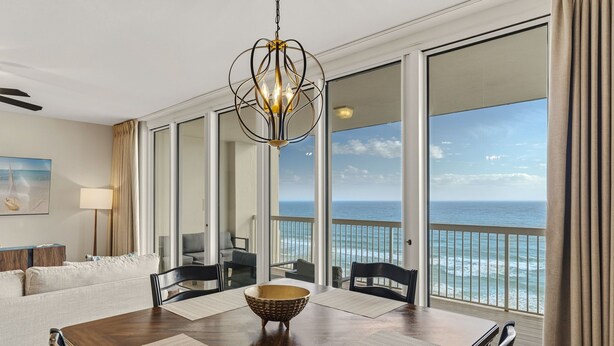 A dining area with a wooden table, chairs, and a central woven bowl. Large windows offer a view of the ocean. - Gulf Front Luxury Condo at Silver Beach Towers (Destin)