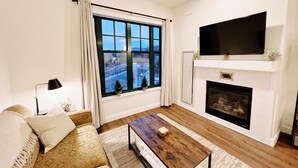 Una sala de estar con sofá, una mesa de centro, chimenea y un amplio ventanal con cortinas.
