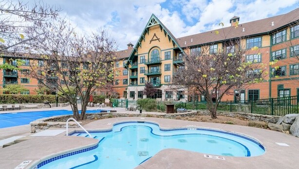 Un hotel con piscina, un edificio grande con un techo triangular característico y un área ajardinada con árboles.