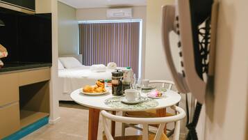Una habitación de hotel compacta con kitchenette, un comedor con mesa lista para dos personas y una cama con colcha blanca.